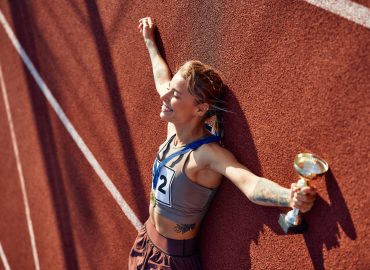 Winner woman in sportswear with number with gold medal for first place and cup lying at stadium celebrating after competition. Sports and  victory concept.